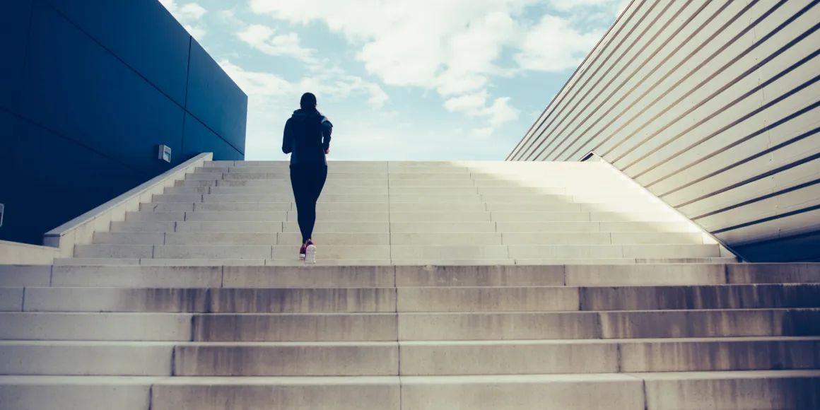 Frau joggt dynamisch eine Treppe hoch an einem sonnigen Tag vor blauem Himmel