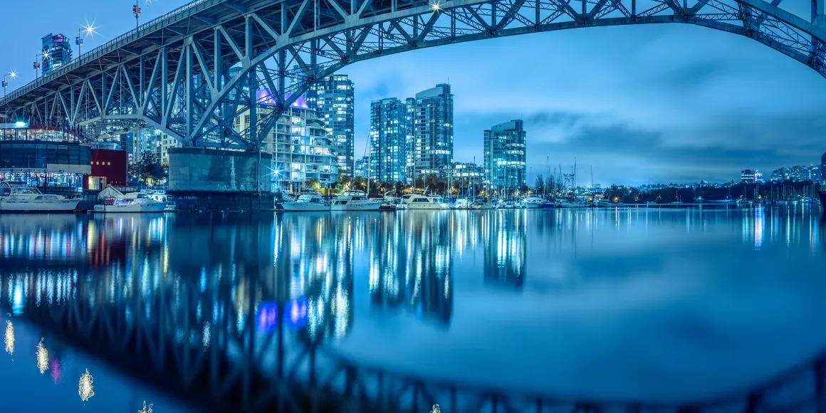 Eine Brücke spiegelt sich bei Nacht im Wasser. Im Hintergrund Yachten & moderne Hochhäuser.